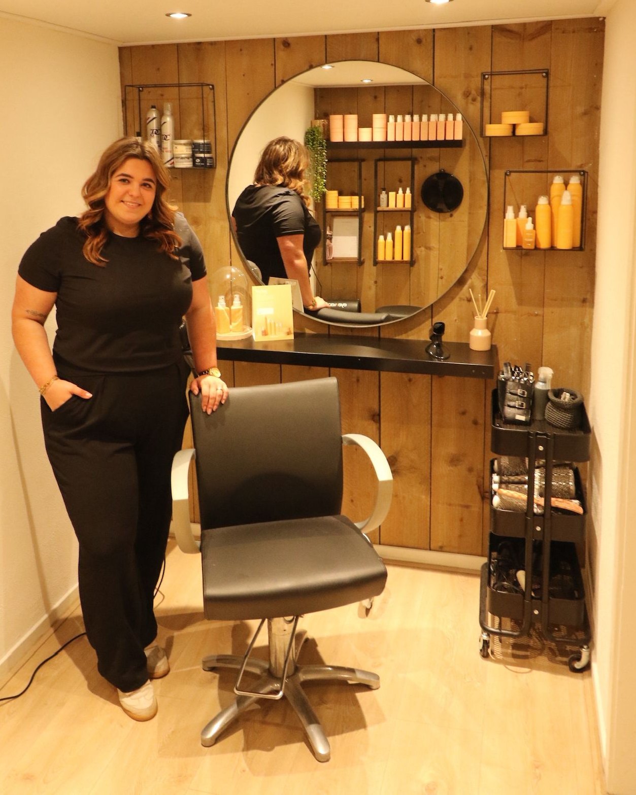 Danique in haar salon in Schijndel, naast de stoel en de ronde spiegel voor de houten wand